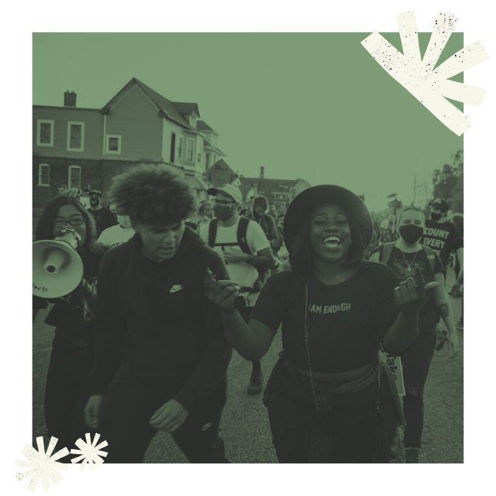 People in protest march walk past houses, chanting and smiling.